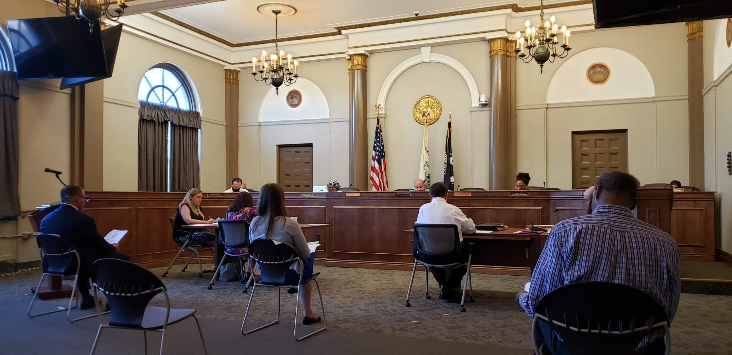Town council chambers with flags and members of the public seated and social distancing.
