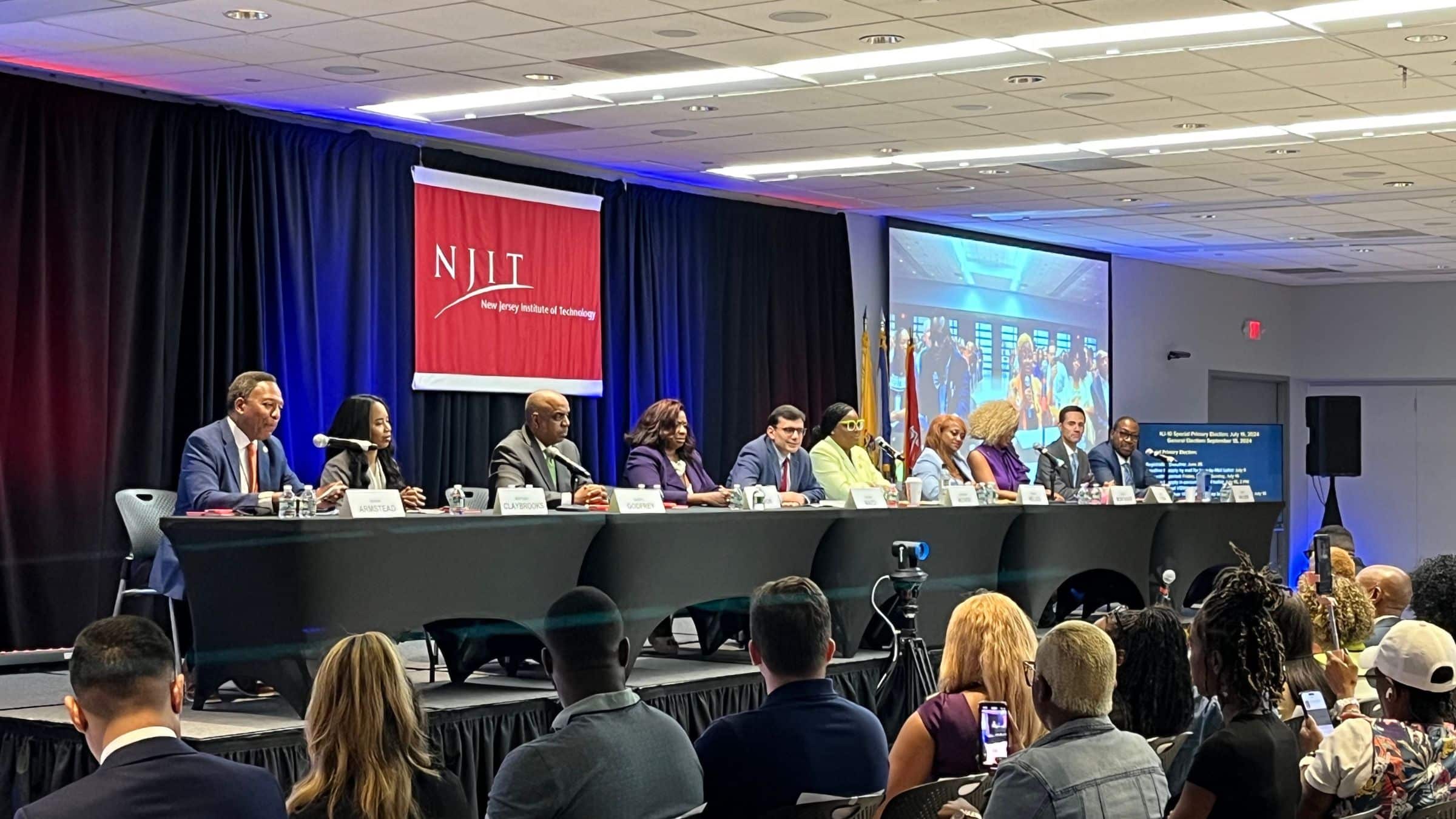 Ten people sit at a long table on stage with a red banner behind them reading "NJIT."