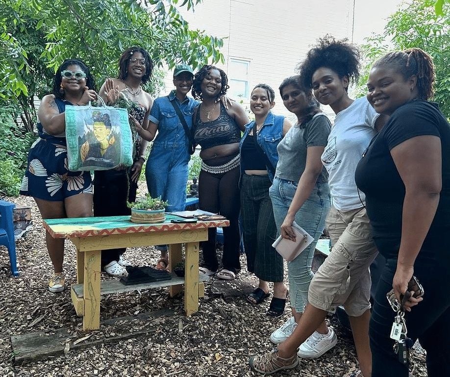A group eight people stand in a garden around a bench with a potted plant on it. One member of the group is holding up a bag with a depiction of the artist Frida Kahlo. 