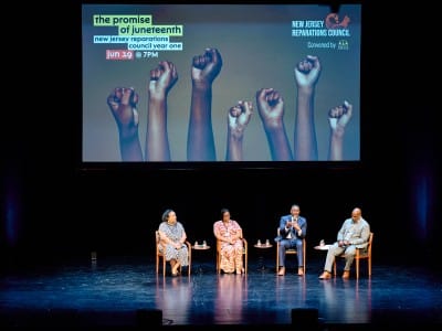 Four people sit in chairs on a stage in discussion below a screen that reads "The Promise of Juneteenth: New Jersey Reparations Council Year One" with the raised fists of people of color pictured.