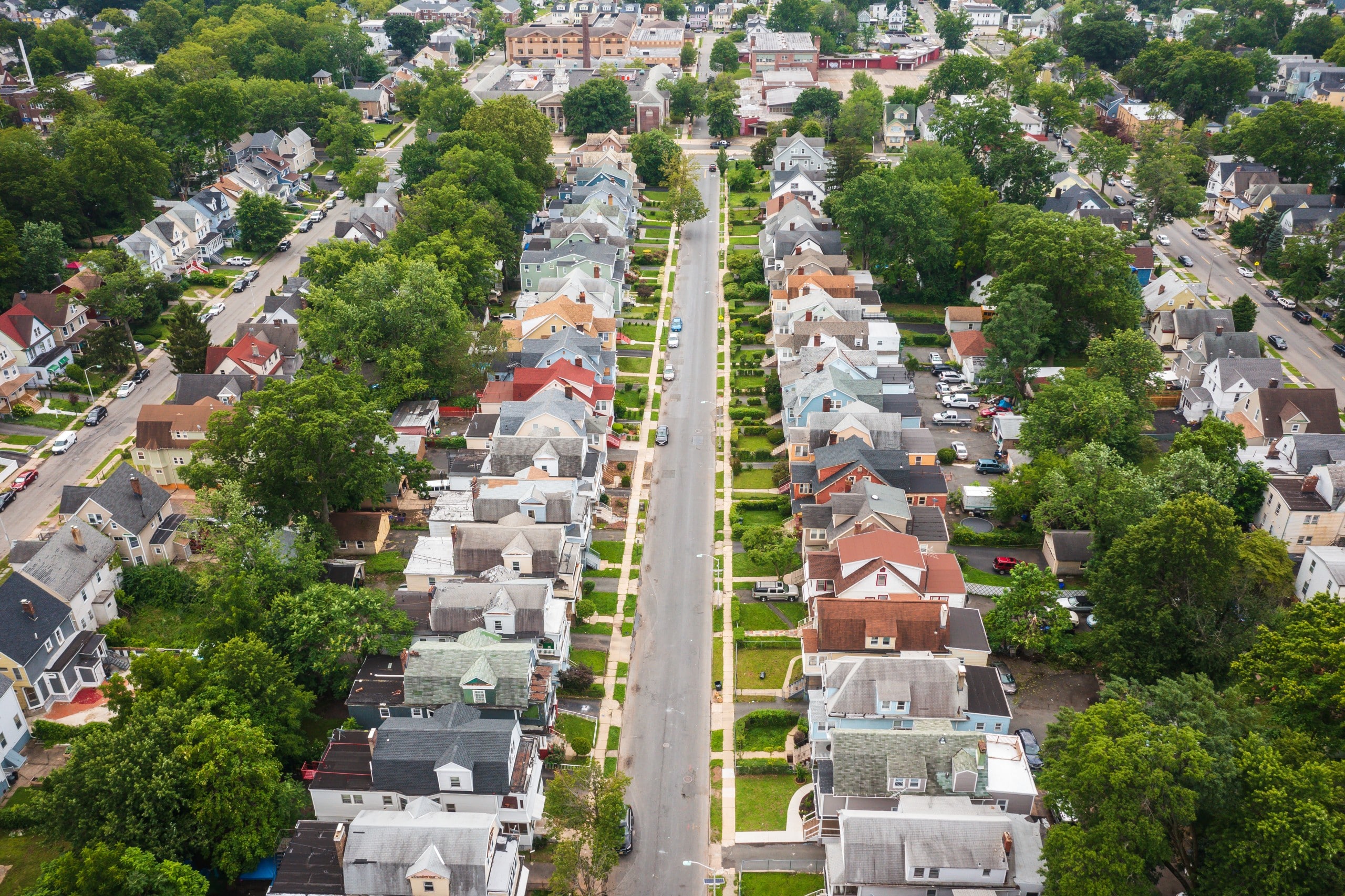 An aerial photo of a neighborhood with a road in running down the middle and multi-family houses on either side, followed by parallel streets and houses on the left and right. There is a school at the end of the street centered in the photo.