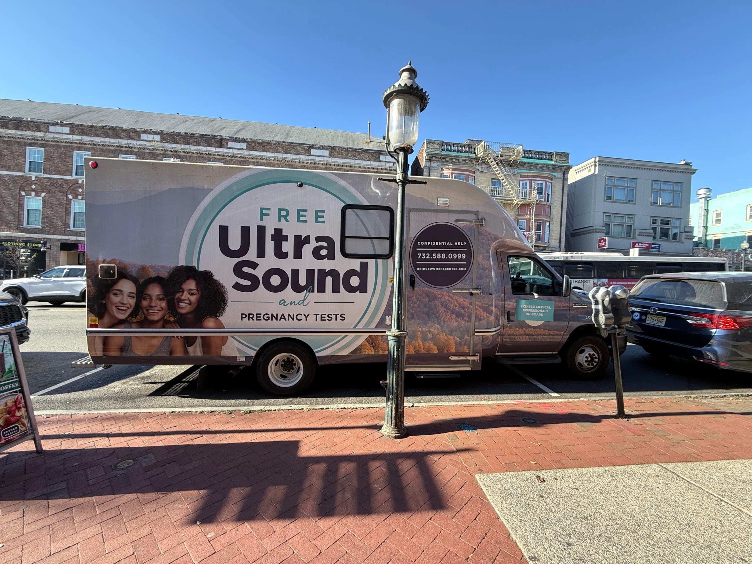A van is parked along a street in front of shops, displaying a large advertisement for free ultrasound and pregnancy tests. The side of the vehicle shows three smiling women alongside bold text reading, "Free Ultrasound and Pregnancy Tests." Additional text offers "Confidential Help" and a phone number (732-588-0999) and website (bridgetowomenscenter.com), suggesting a service related to pregnancy and women’s health. The surrounding area includes a lamp post, a parking meter, and nearby vehicles against a backdrop of storefronts and urban buildings.