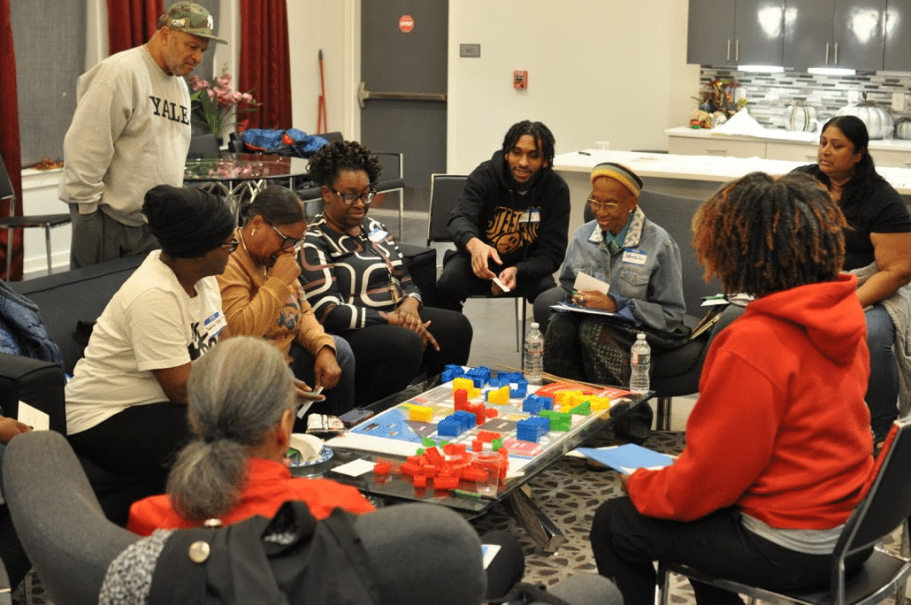 A group of people sit and stand around coffee table where a map is laid flat and topped with blocks of different colors and heights.