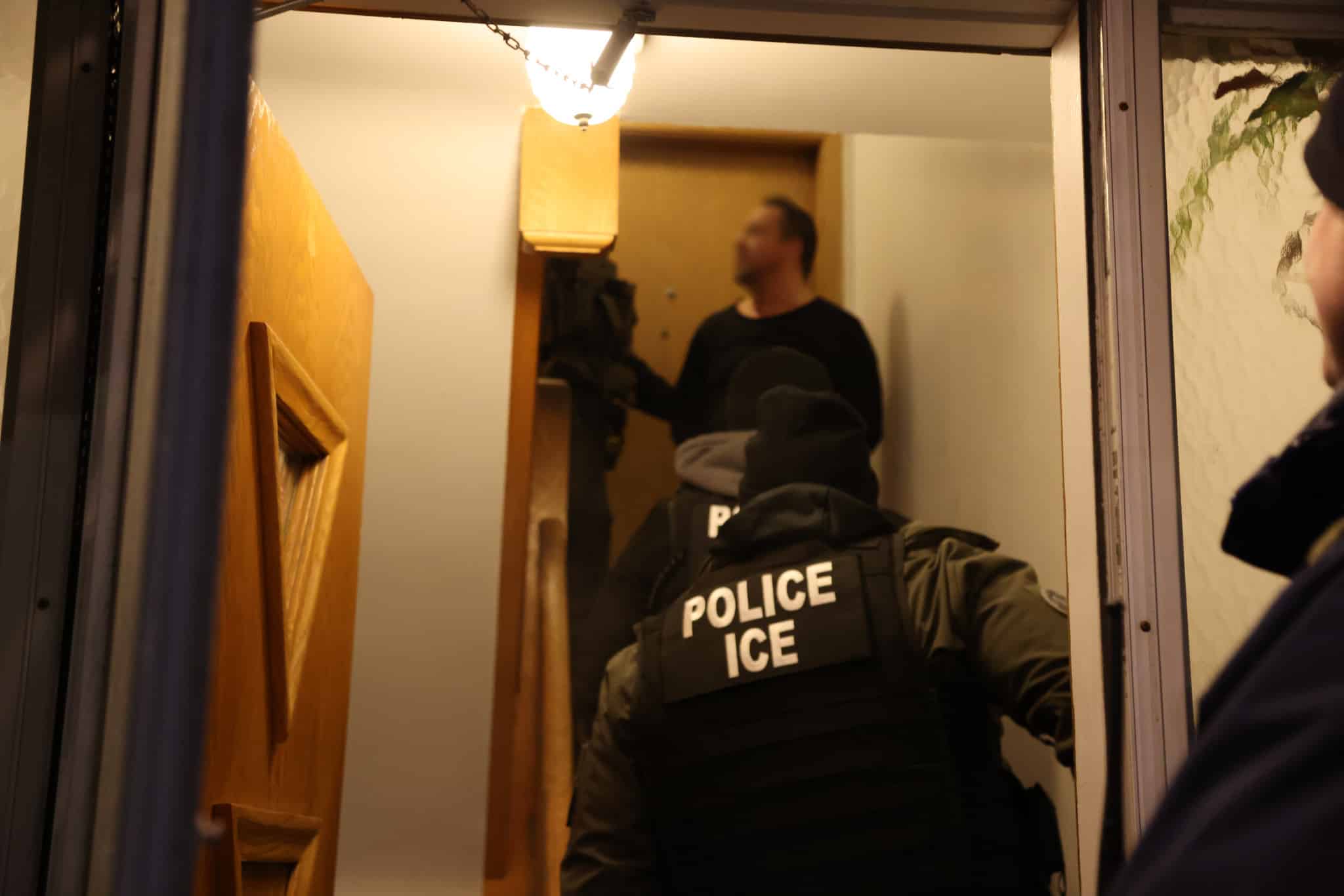 A team of ICE (Immigration and Customs Enforcement) officers in tactical gear enter a home, confronting a man standing at the top of a staircase. The officers wear bulletproof vests labeled "POLICE ICE," and one appears to be carrying equipment. The man, dressed in a black shirt, has a blurred face. The setting is dimly lit, with a wooden front door ajar and a hallway visible inside.